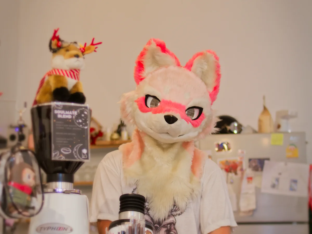 Makarm in a pink and white fursuit behind a café counter